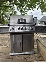 Front view of the stainless steel grill on wooden deck showing double doors, control panel with four knobs and lid closed.