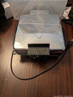 Closed waffle maker showing stainless steel top, black front handle, and attached power cord.