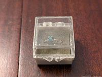 Child's ring with blue square-cut blue stone displayed in a small white plastic box with clear hinged lid, closed box view