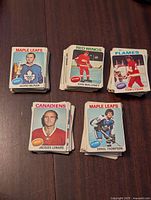 Photo showing multiple stacks of 1975-76 O-Pee-Chee hockey cards featuring players Gord McRae, Dan Maloney, Tom Lysiak, Jacques Lemaire, Errol Thompson from teams Maple Leafs, Red Wings, Flames, Canadiens.