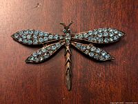 Front of the dragonfly brooch showing detailed metalwork and bright blue stones on wings and body against a wooden background.