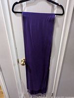Purple cashmere scarf with fringe edges hanging on a black hanger against a white door, showing full length and fringe detail.
