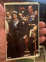 Close-up of postcard showing Queen Elizabeth II in black dress and tiara, standing beside Prince Philip in ceremonial uniform, held in hand