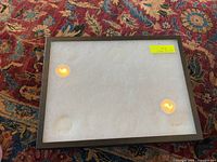Photo of one Ricker mounting glass display case with glass top and dark frame, lying flat on a multicolored patterned carpet. Case has a white interior backing and a yellow price sticker on glass.
