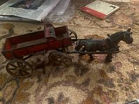 Side view of antique cast iron horse and red wagon toy on patterned carpet, showing worn original paint and metal wheels.