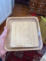 Photo of the bamboo charcuterie board held in hand showing front face with square shape and raised edges, still sealed in plastic wrapping.
