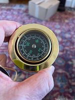 Close-up photo of solid brass compass with black dial face showing degree markings and compass directions, held in hand.
