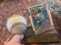 Photo shows the autographed Cal Ripken Jr. baseball in a protective plastic case beside the autographed 1990 Topps Orioles baseball card in a clear holder displayed on a wooden base.