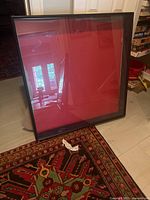 Photo depicts a large square shadow box frame with a black painted wood border and red matting inside. The frame is positioned on a floor next to a door with some household items visible in the background.