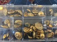 Clear top-down image of compartmentalized case containing approximately 80 brass military buttons, mostly naval with anchor designs.
