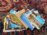 Lot of approximately 85 postcards arranged in a pile on a patterned carpet. Colors and sizes vary, with visible images of buildings and cityscapes.