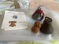 Photo showing a small handwritten note with Liberty Bell sketch, miniature Liberty Bell gold-colored pin, wooden carved bell with eagle design, metal small bell with loop handle, and red metallic clamp bell frame.