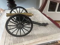 Side view of a miniature model cannon clearly showing black iron carriage and brass barrel, placed on newspaper.