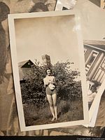 Photo showing young woman in swimsuit standing barefoot outdoors near a chimney and house, black and white photo printed on matte paper.