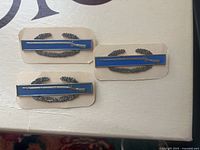 Front view of three US Army blue enamel combat infantry bars with silver rifle design, mounted on cards.