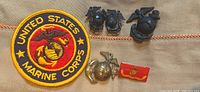 Top view of five US Marine pins on cloth with one circular Marine Corps patch showing embroidered red and yellow emblem and text.