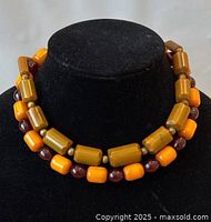 Two Bohemian amber Bakelite necklaces displayed on a black bust. One necklace has large caramel amber cylindrical beads with golden spacer beads; the other has alternating bright orange rectangular and small dark amber round beads.