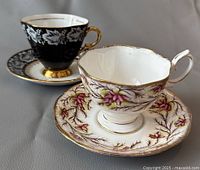 Two English fine bone china teacups with matching saucers, one floral pink and purple with gold trim, other black with white floral pattern and gold trim