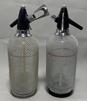 Front view of two 1970s soda syphon bottles with black tops and silver wire mesh covering glass bodies.
