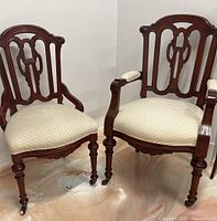 Two vintage solid wood armchairs shown together, one armchair with armrests and one without. Both have upholstered cream seats with small dot patterns and detailed wood backs and legs with caster wheels.