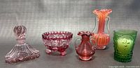 Five vintage art glass pieces displayed together on a gray background: pink perfume bottle, pink footed bowl, small pink ruffled vase, orange-pink swirled vase, green face tumbler.