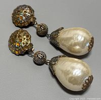 Close-up of two Art Deco style silver-tone dangle clip on earrings with detailed filigree and teardrop faux pearls positioned side-by-side on gray background