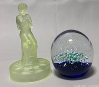 Photo showing the uranium glass Art Deco lady figurine and the round art glass paperweight side by side on a white surface for scale and detail comparison.