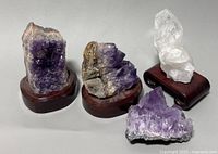 Four natural crystals including three purple amethyst clusters and one clear quartz crystal, all mounted on polished wooden stands.