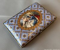 Top angled view showing rectangular shape, classical love-themed artwork on lid, blue and gold geometric patterned background.