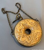 Photo of front of circular vintage powder flask with detailed nude figure relief art, attached chain, and metal spout with lever.