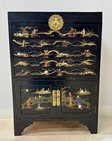 Front view of the black lacquered cabinet chest with floral drawer decorations and hand painted figures on the doors, showing brass hardware.