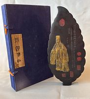 The open blue storage box with the black leaf-shaped ink stick standing beside it, showing intricate gold relief figure and red seal stamps.