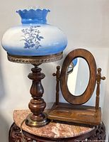 Vintage hurricane lamp with blue and white floral glass globe and wooden base next to oval wooden framed tabletop mirror on carved wooden base.
