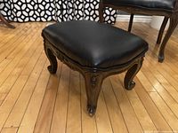 Front angled view showing the carved mahogany legs and black leather upholstered seat of the ottoman, placed on a wood floor with a patterned background.