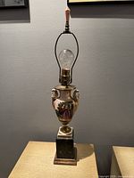Full view of antique French porcelain painted table lamp with detailed painted scene, urn-shaped body, and bulb installed.