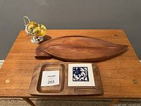 Three assorted items on a wooden table: silver-plated ornate pitcher, wooden oval tray, wooden rectangular tray with tile insert