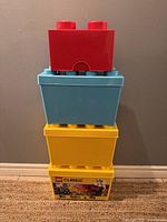 Stacked view of four LEGO storage containers: red brick-shaped on top, blue square, yellow square, and the yellow Classic container with lid at the bottom.