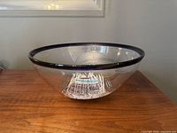 Clear art glass bowl with dark rim and intricate colored designs inside, photographed on a wooden surface under ambient indoor lighting.
