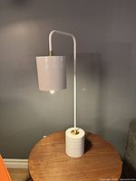 Modern metal table lamp with white cylindrical base and shade on wooden surface against gray wall; lamp is lit in photo.