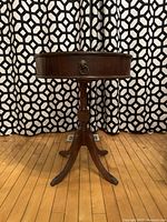 Full front view of vintage round side table showing overall shape, single drawer, and tripod base against patterned curtain backdrop.