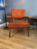 Front view of orange upholstered mid century armchair with wooden legs and armrests.