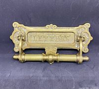 Front view of brass antique door knocker with attached mail slot showing lettering and ornate edges.