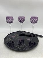 Front view of three purple Bohemian cut crystal wine glasses arranged behind black Mikasa Ebony Meadow floral ceramic cake plate and ceramic cake server knife.