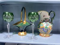 Front view of two green Bohemian cut crystal wine glasses flanking a green and gold glass basket and a chicken-themed ceramic pitcher with green handle.