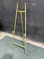 Full view of the brass easel standing upright against a dark wall on a gray carpet.