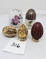 Photo shows five decorative eggs and holders including a white ceramic holder with roses, a gold filigree egg, a red cinnabar egg with floral carvings, a pysanka style egg with chick holder, and a bird painted egg.