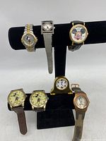 Full group of seven Disney Mickey Mouse wristwatches displayed on a black velvet stand and one small desk clock.