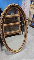 Full view of the large oval mirror leaning against a wall, showing entire frame and reflective surface.
