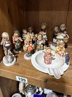 Full view of nine Hummel ceramic figurines displayed on wooden shelf, including boy and girl figures and porcelain ashtray with figurines mounted.