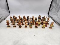 Full view of 30 Hummel porcelain figurines in soft pastel colors depicting children in various poses and traditional clothing on bases.